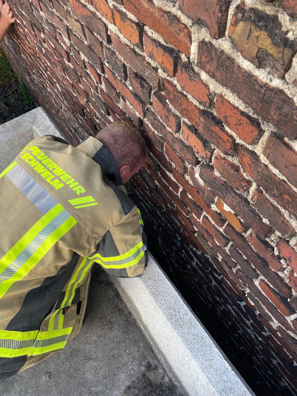 Ein Feuerwehrmann von der Feuerwehr Schwelm beugt sich vor eine Ziegelmauer, um einen Spalt zu überprüfen. Der Mann trägt eine feuerfeste Jacke mit reflektierenden Streifen, und der Boden ist betoniert.