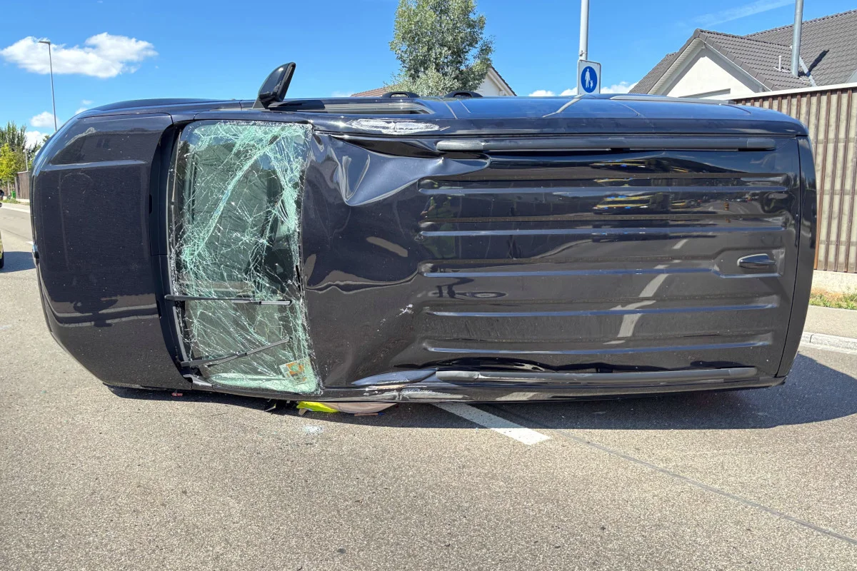Ein Auto liegt seitlich auf einer Straße. Die Windschutzscheibe ist stark beschädigt, und es scheint bei Tageslicht aufgenommen worden zu sein.