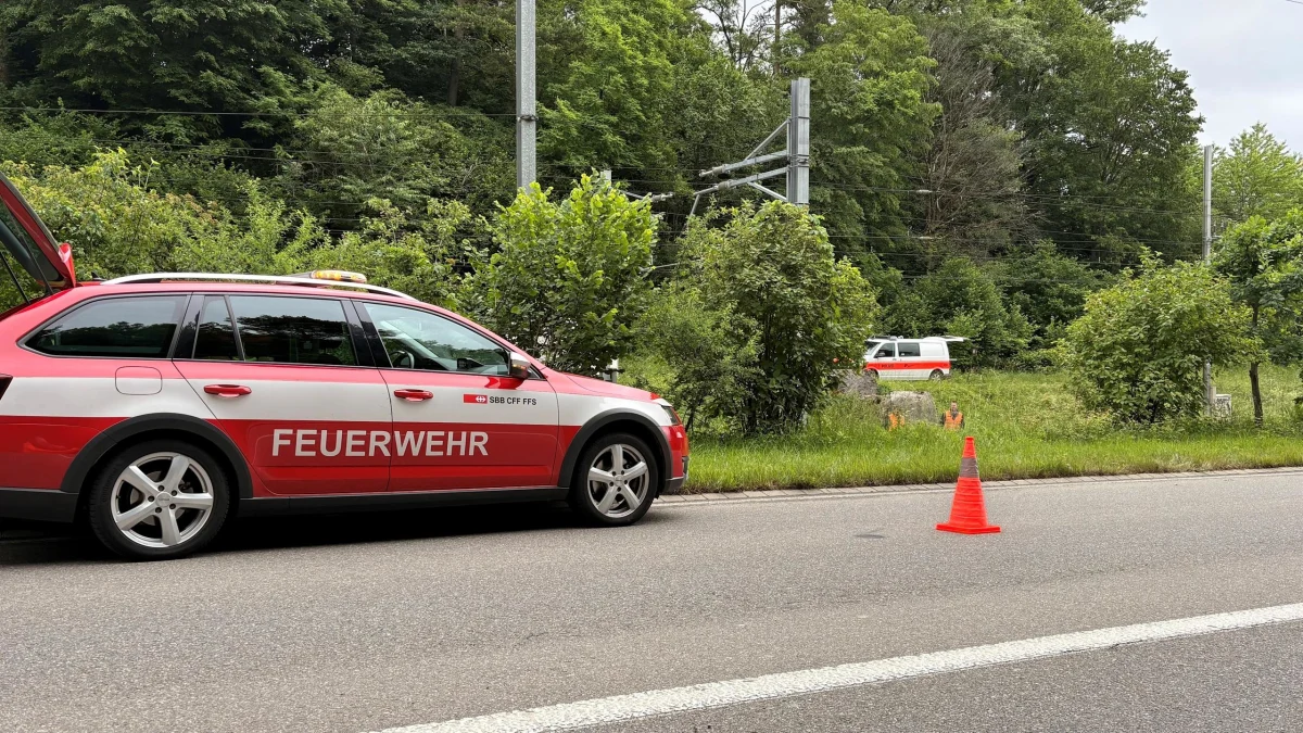 Ein rotes Feuerwehrfahrzeug mit offenen Türen steht auf einer Straße neben einer Bahnstrecke. Es sind auch Verkehrsleitkegel zu sehen.