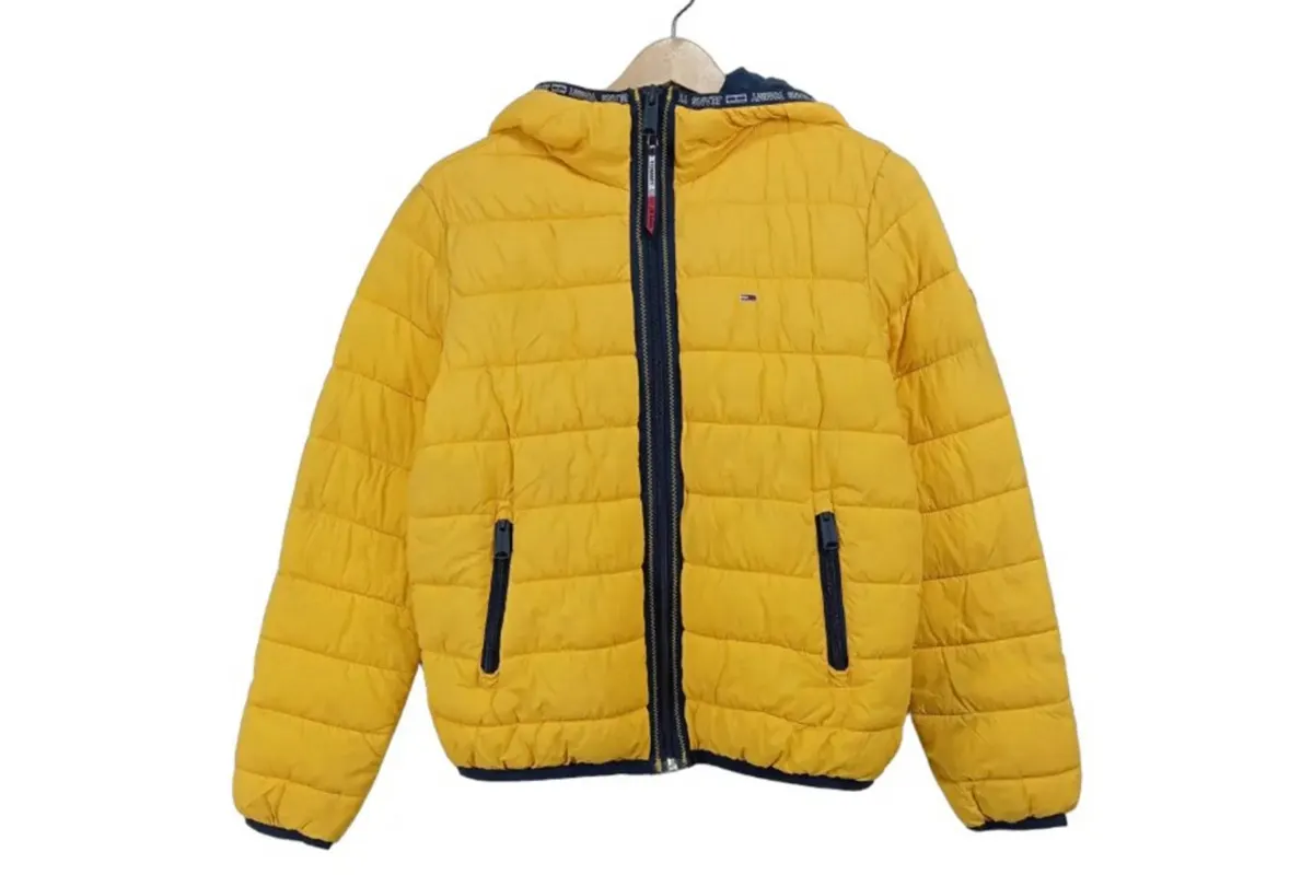 Eine leuchtend gelbe gesteppte Jacke hängt auf einem hölzernen Kleiderbügel. Die Jacke hat schwarze Reißverschlüsse und einen schmalen schwarzen Rand an der Kapuze.
