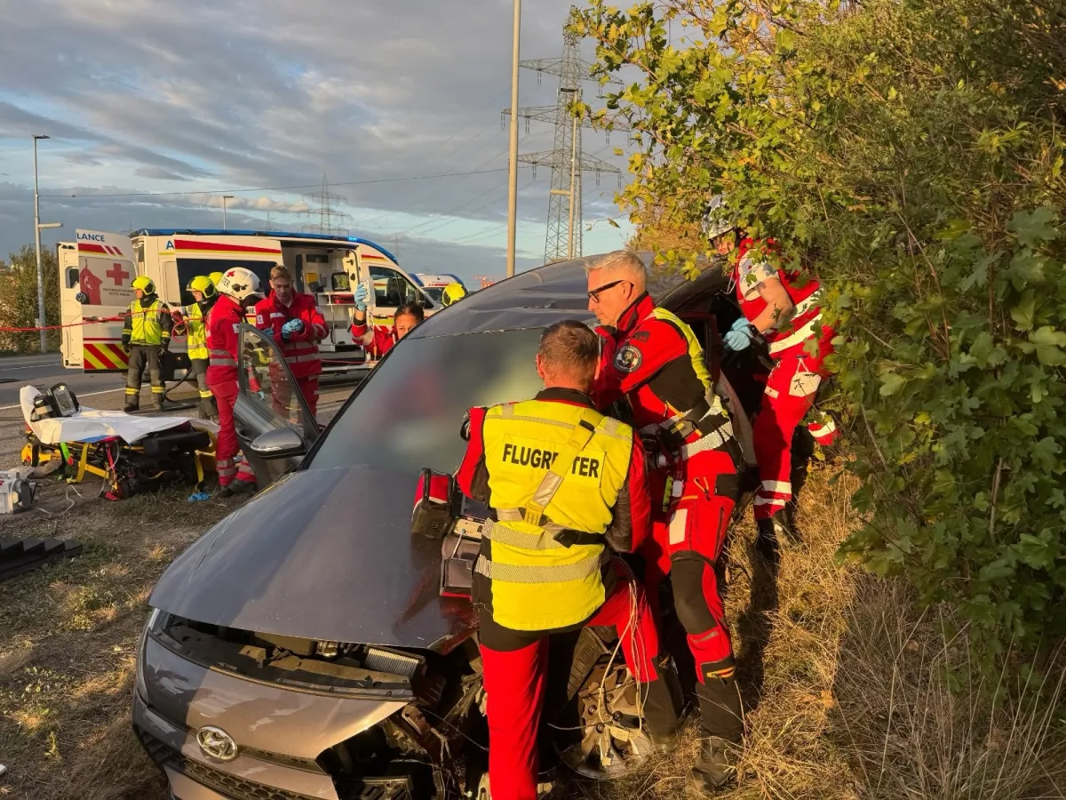Mehrere Rettungskräfte sind dabei, eine Person nach einem Unfall aus einem Auto zu befreien. Im Hintergrund ist ein Rettungswagen zu sehen.