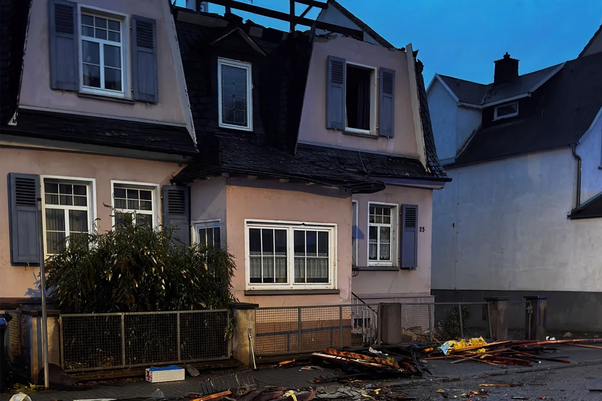 Ein zweistöckiges Haus mit schweren Schäden am Dach und Trümmern davor. Die Fenster sind intakt, aber das Dach ist teilweise eingestürzt.