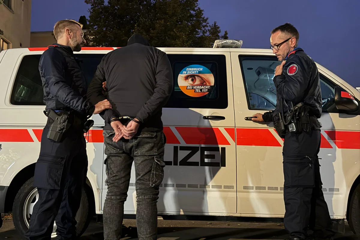 Zwei Polizisten nehmen einen Mann in Handschellen vor einem weiß-roten Polizeifahrzeug fest. Es ist Abend.