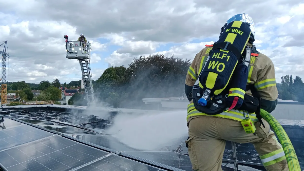Ein Feuerwehrmann in voller Schutzausrüstung spritzt Wasser auf brennende Solarmodule auf einem Dach. Im Hintergrund ein Feuerwehrmann auf einer Leiterbühne.