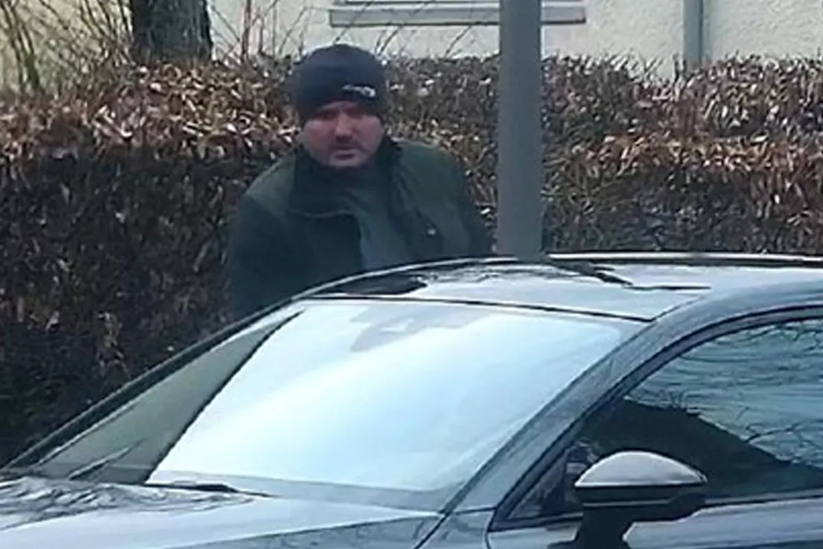 Ein Mann mit dunkler Mütze und dunkler Kleidung steht neben einem schwarzen Auto. Im Hintergrund sind Büsche zu sehen.