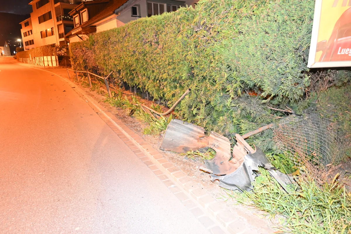 Bei Nacht zeigt das Bild einen zerstörten Zaun und beschädigte Pflanzen an einer Straße. Die Straße ist beleuchtet, und es sind Trümmer erkennbar.