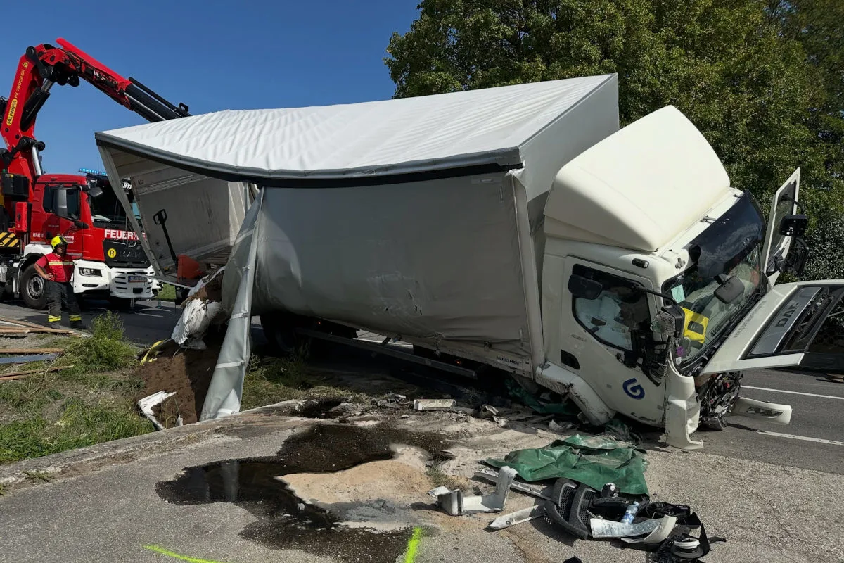 Ein beschädigter Lkw liegt mit offenem Führerhaus am Straßenrand, während ein Feuerwehrfahrzeug im Hintergrund sichtbar ist.