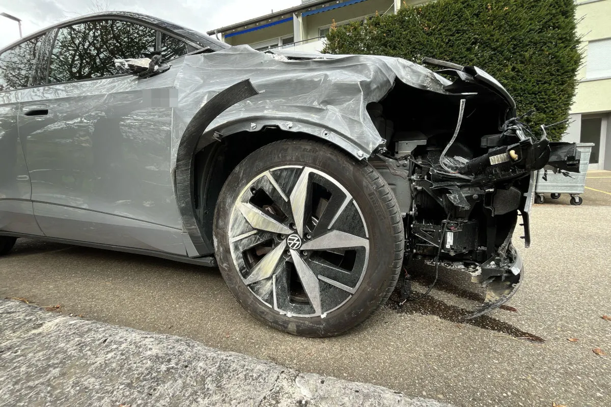 Ein graues Auto zeigt erhebliche Schäden an der Vorderseite, der Stoßfänger ist eingedrückt und Teile liegen frei. Im Hintergrund sind Büsche und ein Gebäude zu sehen.