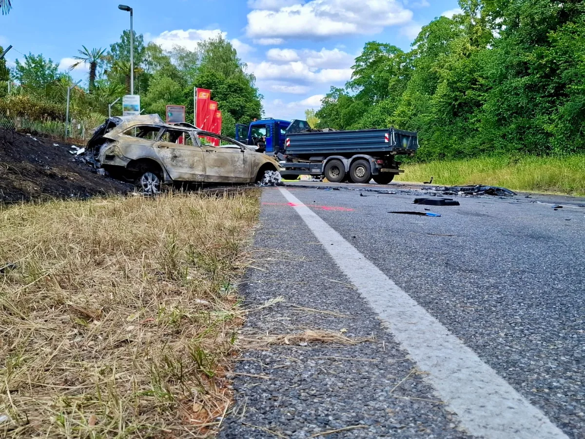 Ein stark beschädigtes, ausgebranntes Auto steht am Straßenrand nahe einer grünen Böschung neben einem Lkw. Trümmer liegen verstreut auf der Straße.