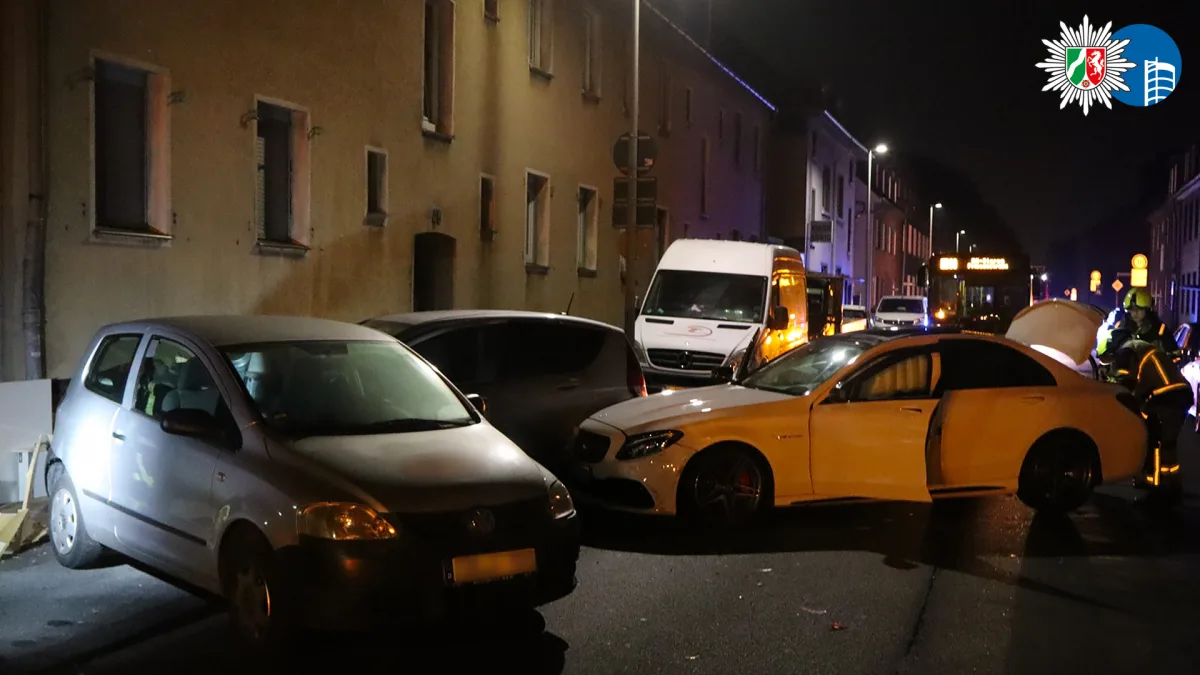 Ein weißer Mercedes steht mit offener Tür quer vor zwei geparkten Autos bei Nacht. Einsatzkräfte sind sichtbar.