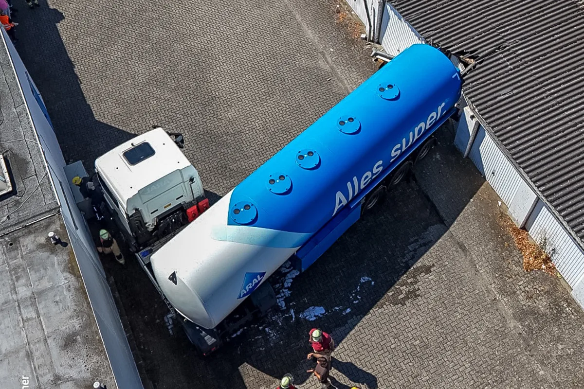 Ein blauer Tanklaster mit der Aufschrift 'Alles super' ist in die Wand einer Halle gefahren. Feuerwehrleute sind vor Ort.