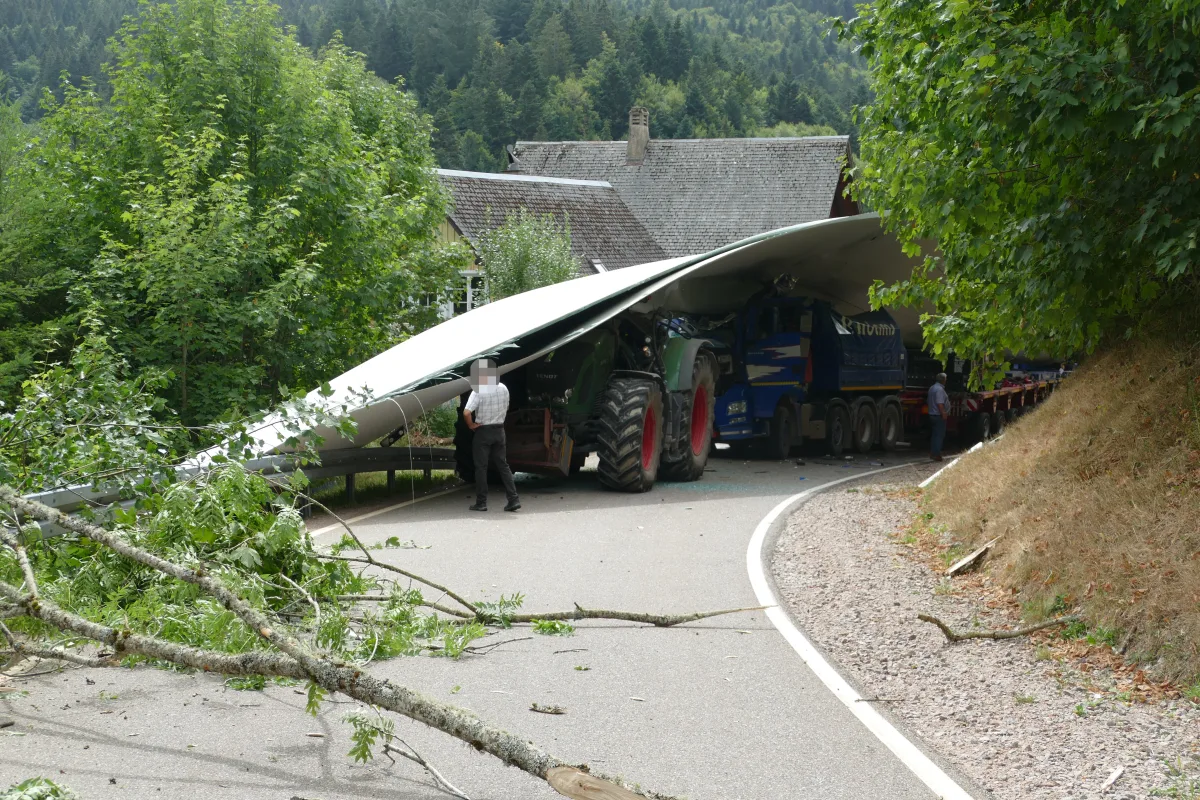 Ein übergroßer Windradflügel auf einem Lastwagen hat sich bei der Durchfahrt um eine Kurve an einem Baum verkeilt. Zwei Personen stehen daneben, um die Situation zu begutachten.