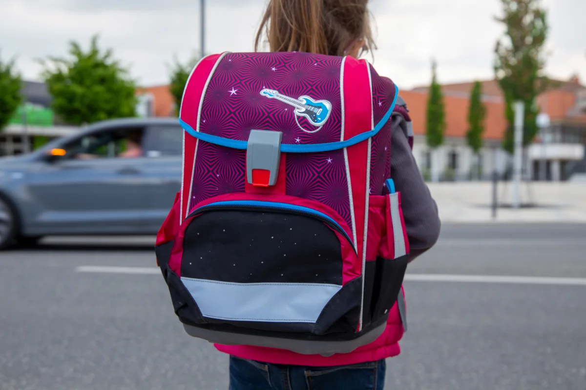 Ein Kind mit einem prägnant rot-blauen Rucksack und Gitarrenmotiv steht mit dem Rücken zur Kamera vor einer Straße. Ein vorbeifahrendes Auto ist im Hintergrund sichtbar.