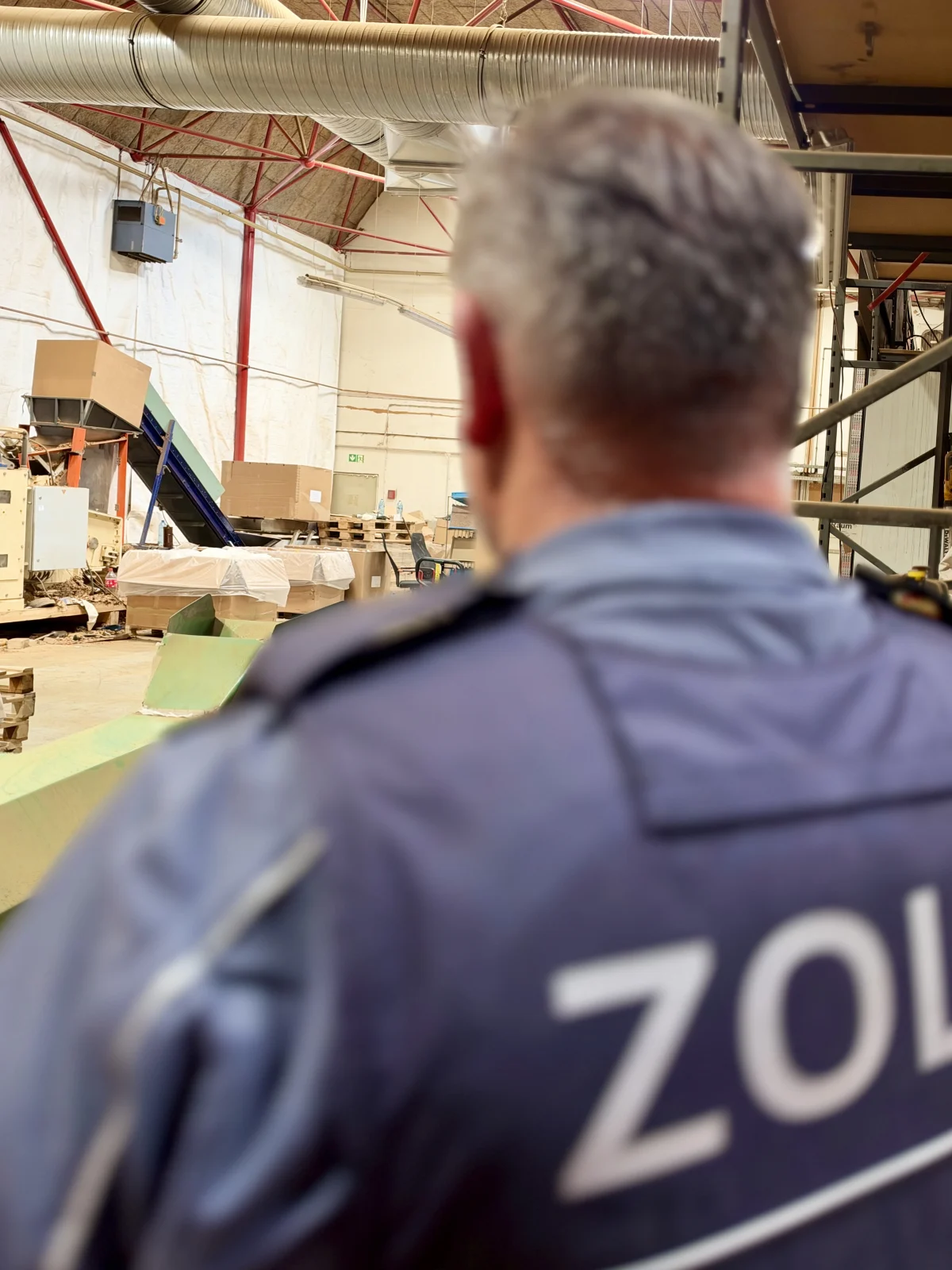 Ein Zollbeamter im Vordergrund überwacht eine Lagerhalle mit Kartons und Industrieanlagen im Hintergrund. Die Szene wirkt aufgeräumt, und die Gebäudeinfrastruktur mit Lüftungsrohren und Stahlträgern ist sichtbar.