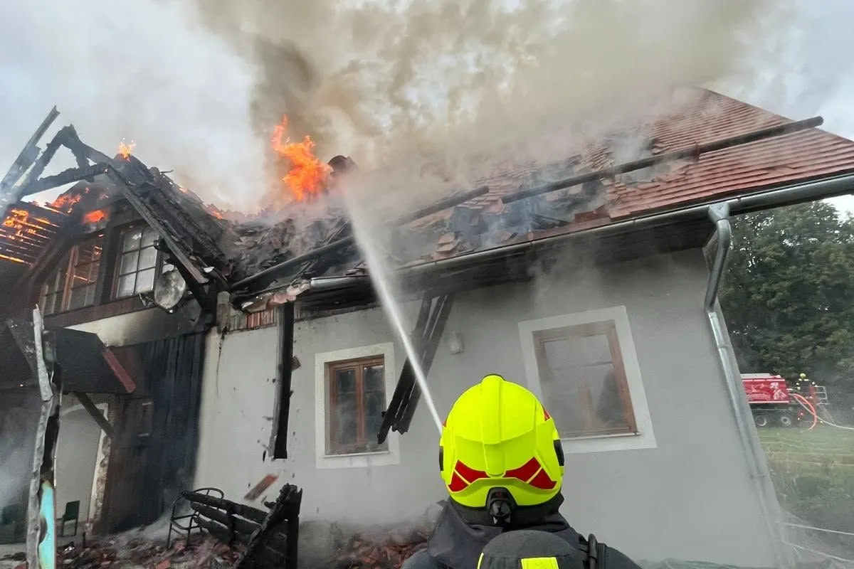 Ein Feuerwehrmann mit gelbem Helm löscht ein brennendes Dach mit einem Wasserschlauch. Flammen und Rauch steigen aus dem Dach auf.
