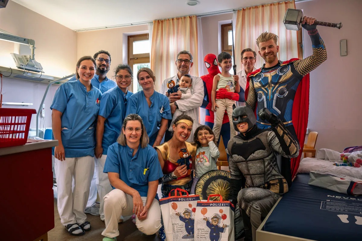 Krankenhauspersonal und mehrere Personen in Superheldenkostümen posieren mit zwei Kindern in einem Raum.