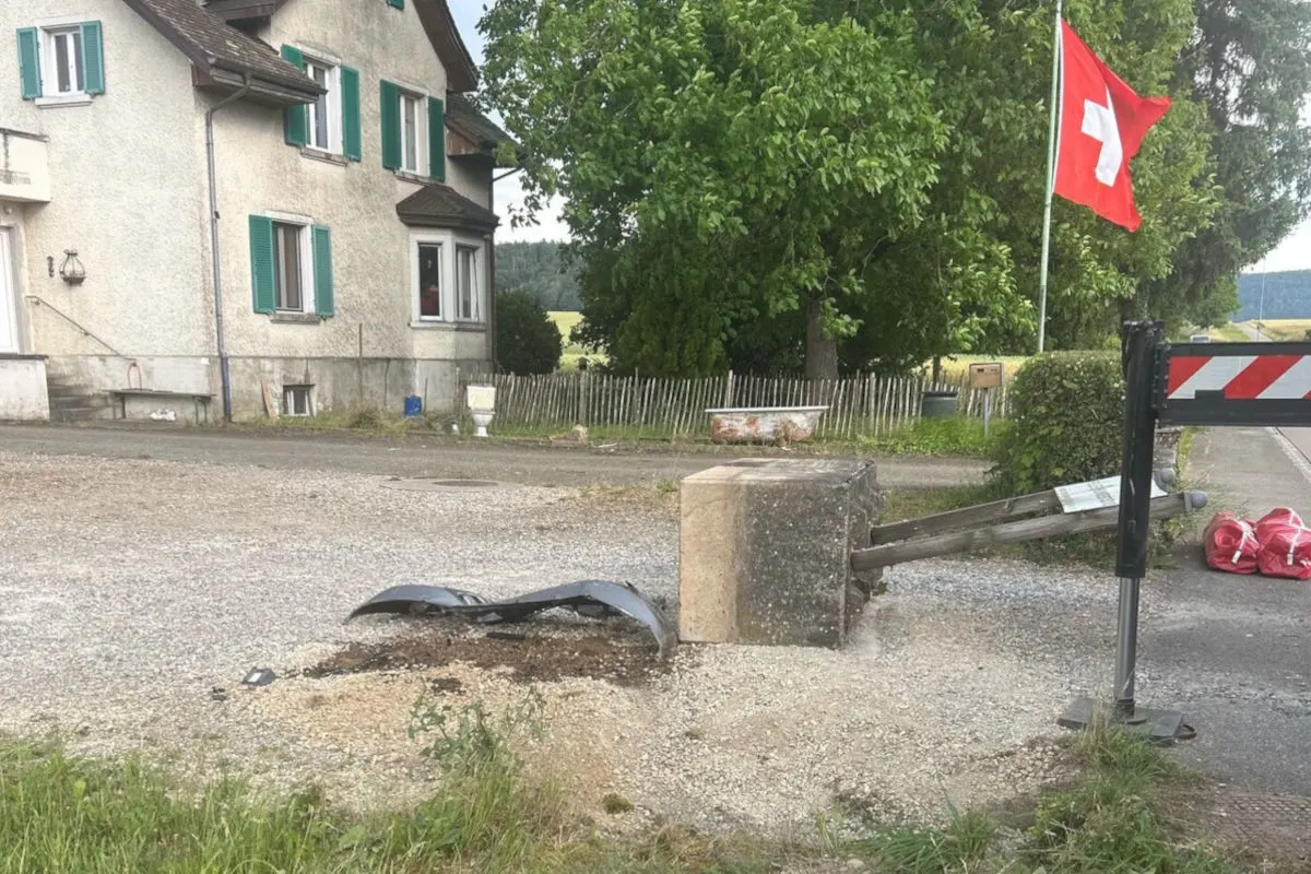 Ein umgestürzter Poller liegt auf einer kiesigen Fläche neben einer Schweizer Flagge und einem alten Haus mit grün-weißen Fensterläden.