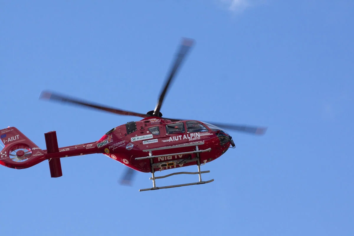Ein roter Rettungshubschrauber von AIUT ALPIN fliegt am blauen Himmel. Der Hubschrauber ist mit mehreren Logos und Schriftzügen versehen.