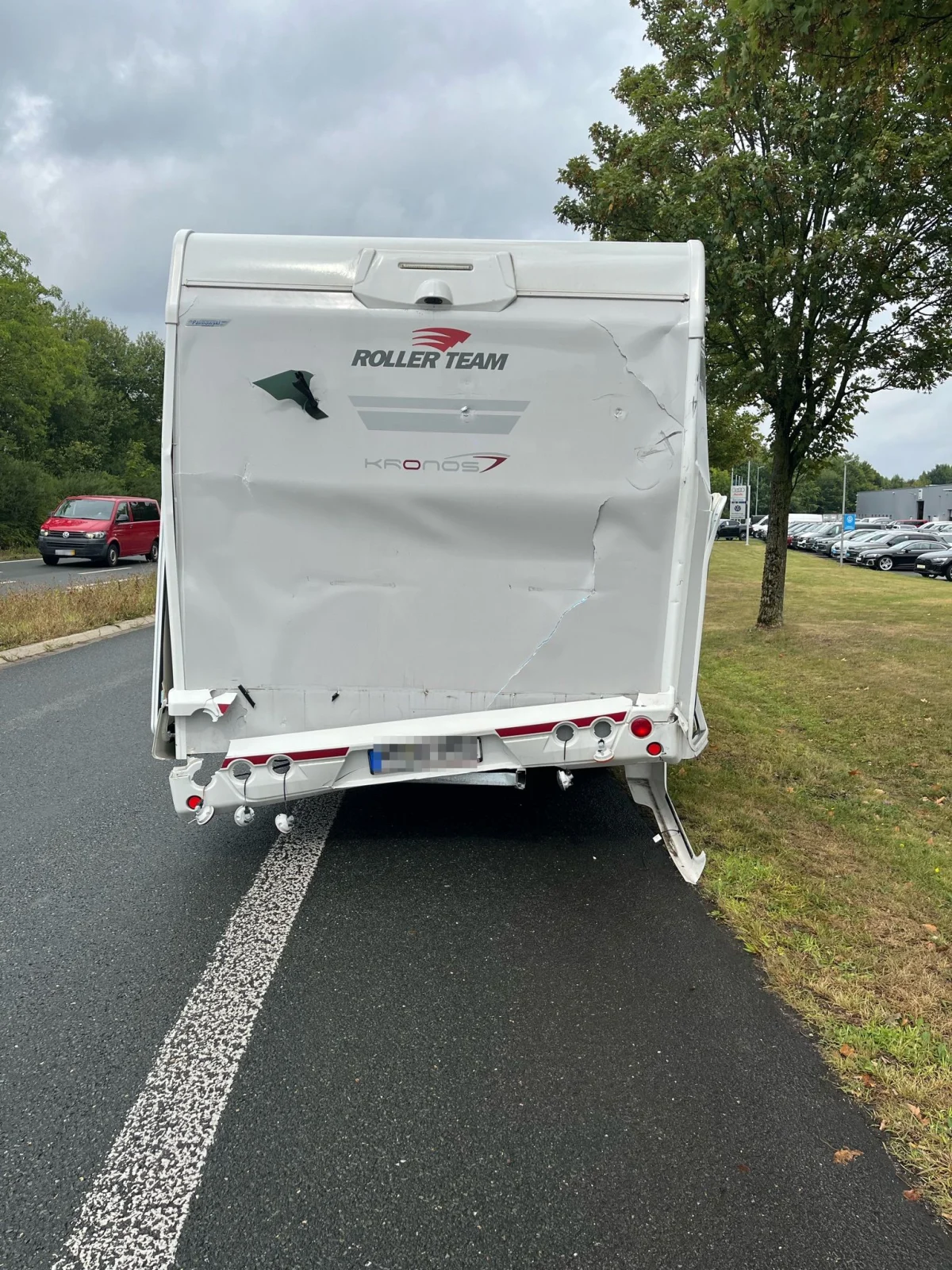 Das Bild zeigt das beschädigte Heck eines Wohnmobils mit Dellen und Kratzern, das am Straßenrand geparkt ist. Ein rotes Fahrzeug ist in der Ferne sichtbar, während Bäume und eine Rasenfläche die Umgebung bilden.