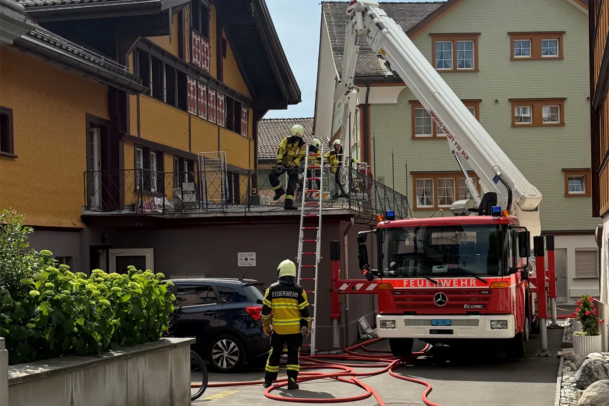 Mehrere Feuerwehrleute arbeiten auf einem Balkon, während ein Feuerwehrfahrzeug mit Leiter im Vordergrund steht. Das Bild zeigt eine städtische Wohnszene bei Tageslicht.