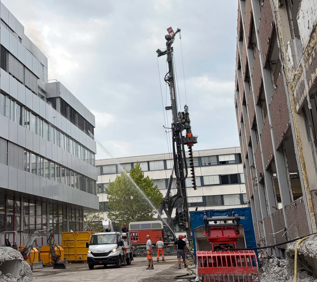 Auf einer Baustelle zwischen Bürogebäuden ist ein großes Bohrgerät zu sehen, umgeben von Bauarbeitern. Ein Lastwagen und andere Maschinen sind ebenfalls vorhanden.