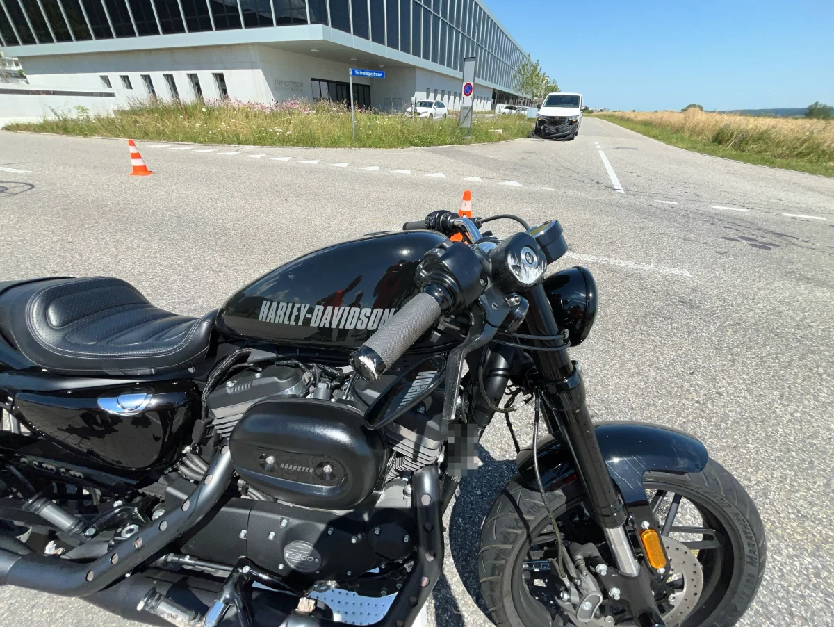 Ein schwarzes Harley-Davidson-Motorrad steht auf einer asphaltierten Straße. Im Hintergrund ist ein modernes Gebäude sowie ein weißer Lieferwagen zu sehen.