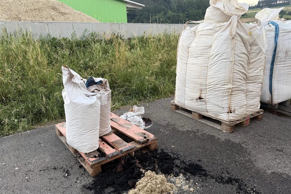 Zwei große weiße Big Bags stehen auf Paletten auf einer Asphaltfläche. Einer der Säcke ist beschädigt und schwarze Substanz liegt daneben auf dem Boden.