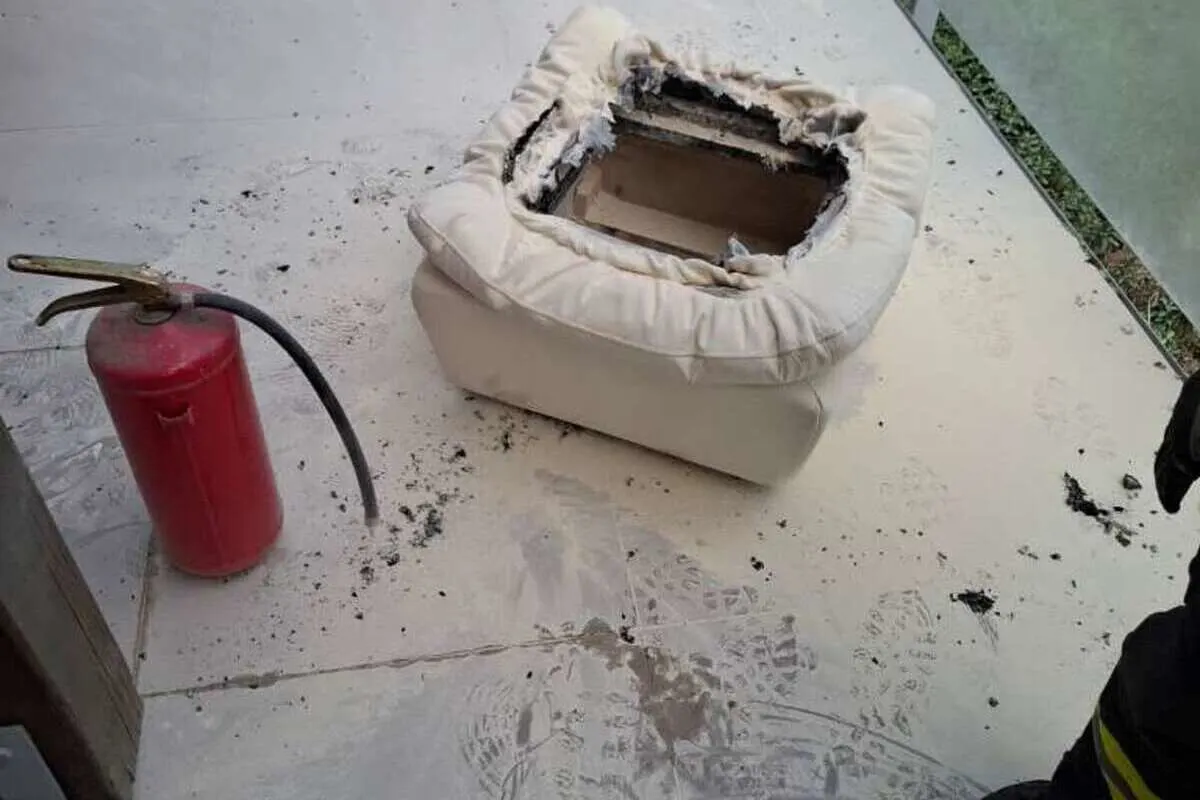 Ein ausgebrannter Sitz liegt auf einem Boden, umgeben von Asche, mit einem roten Feuerlöscher daneben. Fußabdrücke sind auf dem Boden sichtbar.