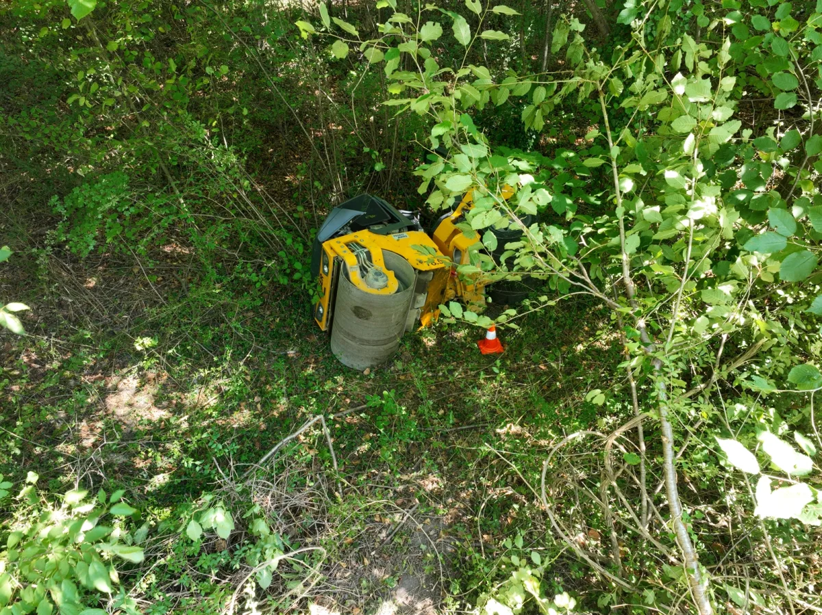 Ein gelb-schwarzes Baugerät ist auf die Seite gekippt und liegt im dichten Gebüsch eines bewaldeten Gebiets.