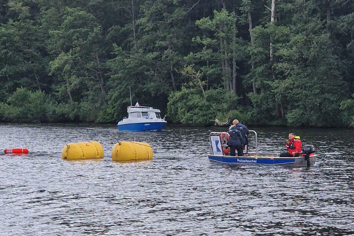 Zwei Personen in Polizeiuniform sind auf einem kleinen Einsatzboot, im Hintergrund sind gelbe Bojen und ein weiteres Schiff zu sehen.