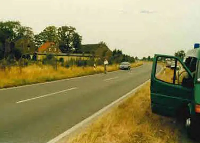 Ein grünes Polizeifahrzeug steht am Straßenrand einer Landstraße, während ein Auto in der Ferne fährt. Die Umgebung ist ländlich mit wenigen Gebäuden im Hintergrund.