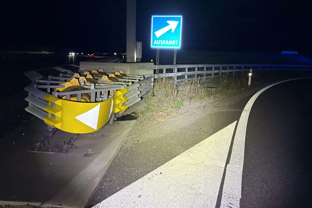 Eine beschädigte Leitplanke ist im Vordergrund einer Autobahnausfahrt zu sehen; ein großes, beleuchtetes Ausfahrtsschild steht daneben.