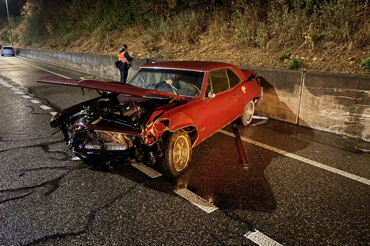 Ein stark beschädigtes rotes Auto steht auf einer Autobahn mit schwer deformierter Front. Eine Person trägt eine Warnweste in der Nähe.