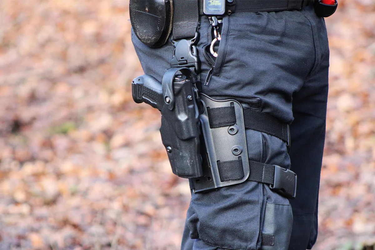Eine Person in dunkler Uniform steht im Freien, sichtbar ist ein Holster am Gürtel, der Wald im Hintergrund ist unscharf.