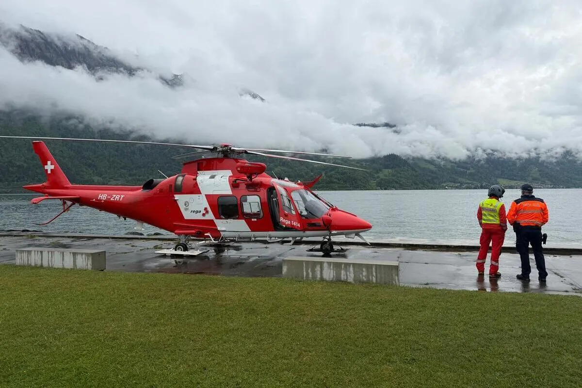Ein roter Helikopter der Schweizer Rega steht auf einem betonierten Bereich am See. Zwei Personen stehen neben dem Hubschrauber, eine in roter Einsatzkleidung, die andere in einer orange-blauen Uniform.