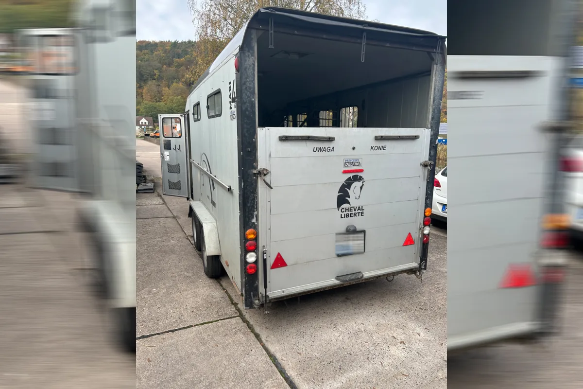 Ein grauer Pferdeanhänger mit der Aufschrift 'Cheval Liberte' steht unverschlossen mit geöffneter Heckklappe auf einem Parkplatz. Neben dem Anhänger sind Teile eines weißen Autos sichtbar.