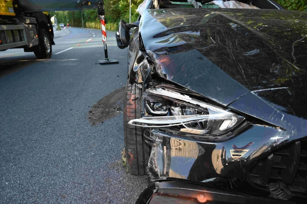 Die Vorderseite eines schwarzen Autos ist stark beschädigt, mit sichtbaren Kratzern und einem kaputten Scheinwerfer. Im Hintergrund ist eine Landstraße zu sehen.