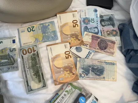 Auf einem Bett liegen verschiedene Banknoten ausgebreitet, darunter Euros, US-Dollar und asiatische Währungen.