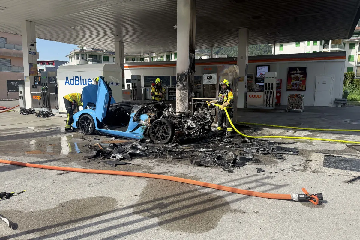 Ein stark beschädigtes, verbranntes Auto steht vor einer Tankstelle. Feuerwehrleute in Schutzkleidung sind mit einem Wasserschlauch vor Ort.