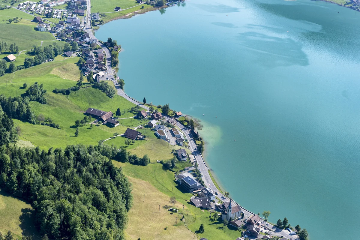 Eine Luftaufnahme zeigt einen klaren blauen See umgeben von einem kleinen Dorf und grünen Feldern. Im Hintergrund erstreckt sich eine Straße.