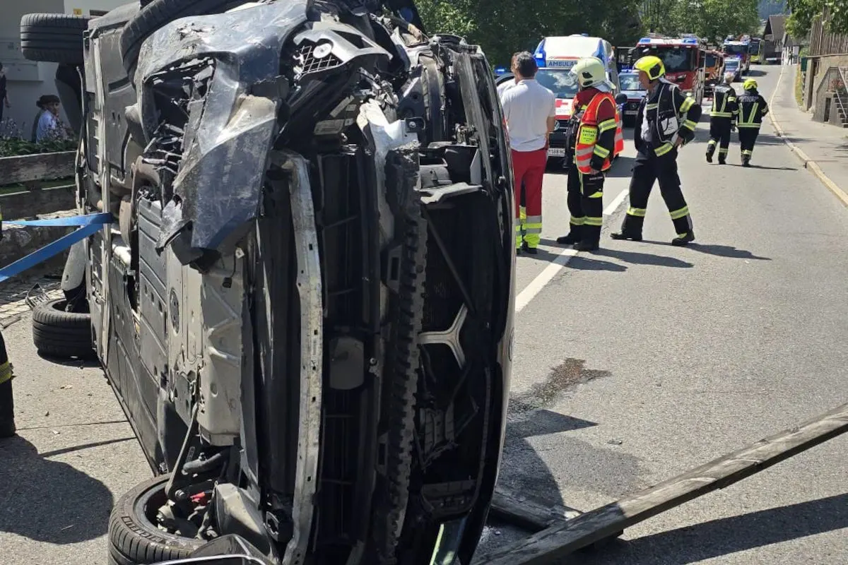 Ein schwer beschädigtes Auto liegt umgestürzt auf der Seite auf einer Landstraße. Daneben stehen Einsatzkräfte.