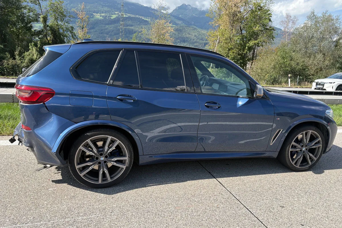 Ein blauer BMW-SUV steht seitlich auf einer Straße. Im Hintergrund sind Berge und Bäume zu sehen.