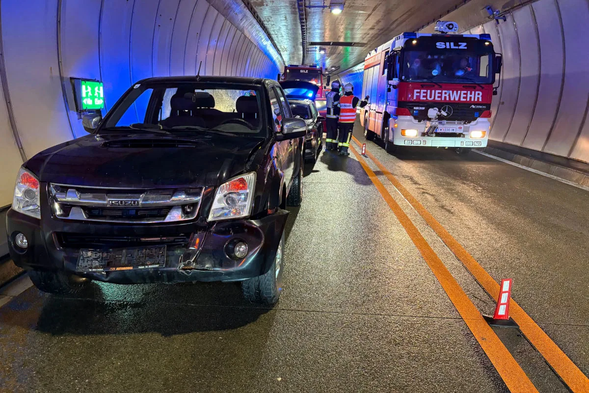 In einem Tunnel steht ein beschädigtes schwarzes Auto mit einer Beule in der Front. Rechts daneben ist ein Feuerwehrfahrzeug mit der Aufschrift 'Silz' zu sehen, umgeben von Rettungskräften.