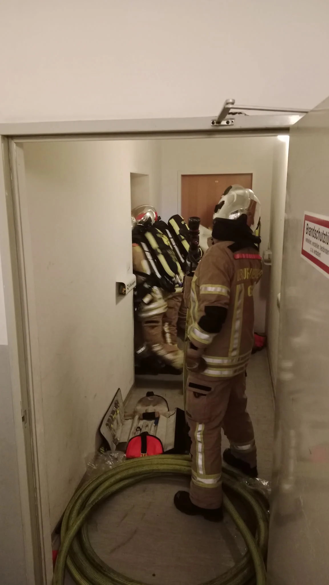Zwei Feuerwehrleute in vollständiger Schutzkleidung und Atemschutzgeräten betreten durch einen engen Flur ein Gebäude für einen Noteinsatz.