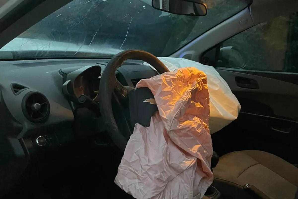 Das Bild zeigt einen aufgeblasenen Airbag im Innenraum eines Fahrzeugs nach einem Unfall. Der Airbag ist sichtbar vor dem Lenkrad, während der Rest des Fahrzeuginnenraums teilweise im Schatten liegt.
