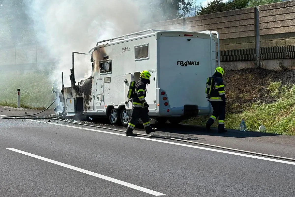 Auf einer Autobahn ist ein Wohnwagen in Brand geraten, während Feuerwehrleute in Schutzanzügen die Szene sichern. Rauch steigt aus dem Fahrzeug auf.
