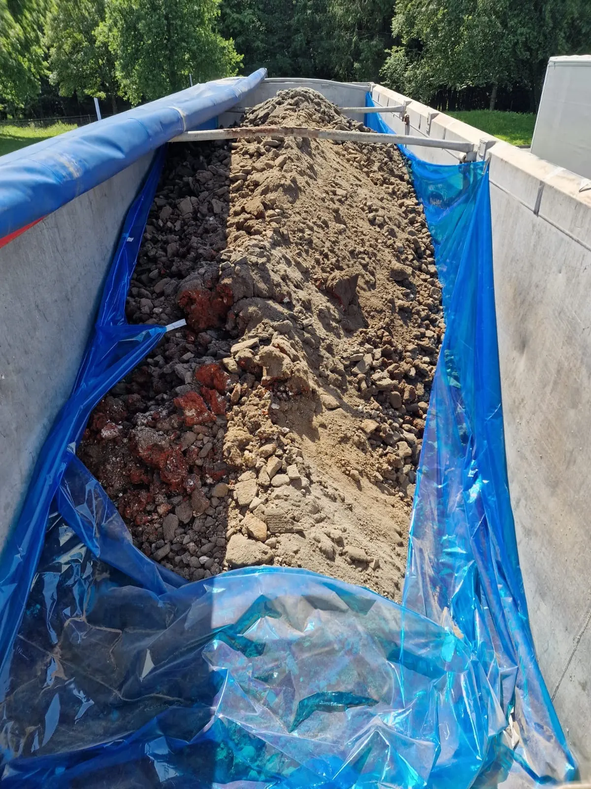 Ein großer Container ist mit Erde und Bauschutt gefüllt. Blaue Planen sind rund um den Container zur Abdeckung angebracht.