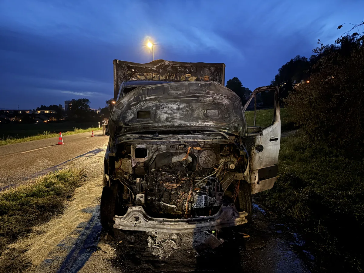 Ein ausgebranntes Fahrzeug steht mit geöffneten Türen am Straßenrand. Hinter dem Fahrzeug sind Verkehrsleitkegel sichtbar, die den Bereich absichern.