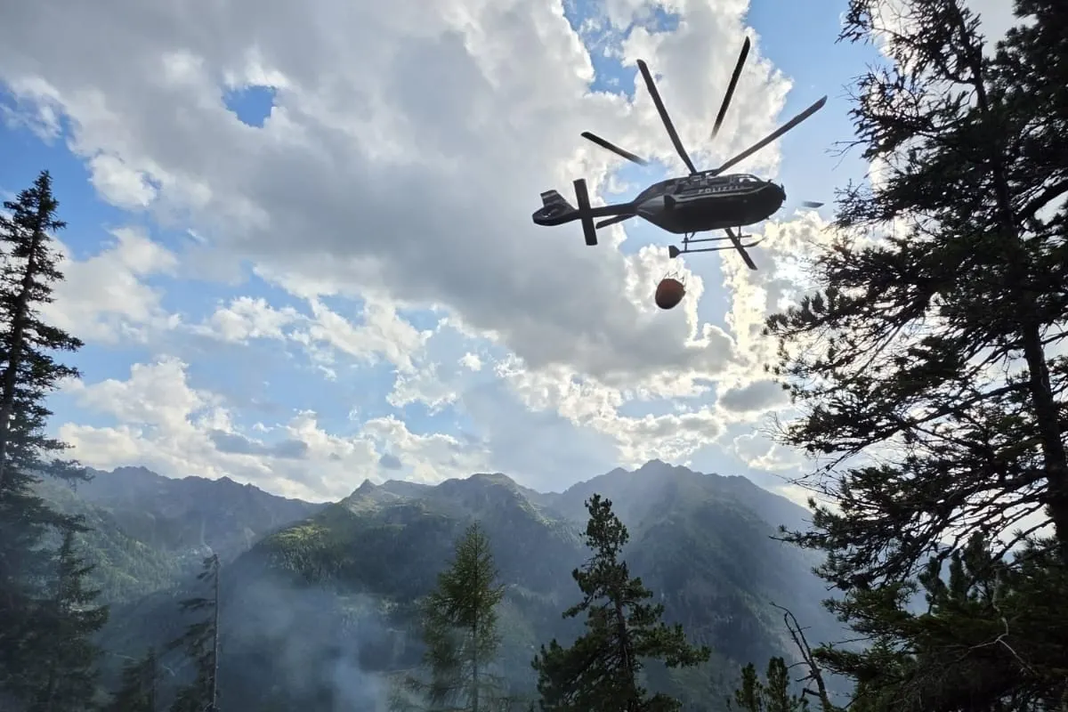 Ein Polizeihubschrauber fliegt mit einem Löschwasserbehälter durch ein bewaldetes Berggebiet. Der Himmel ist teils bewölkt.