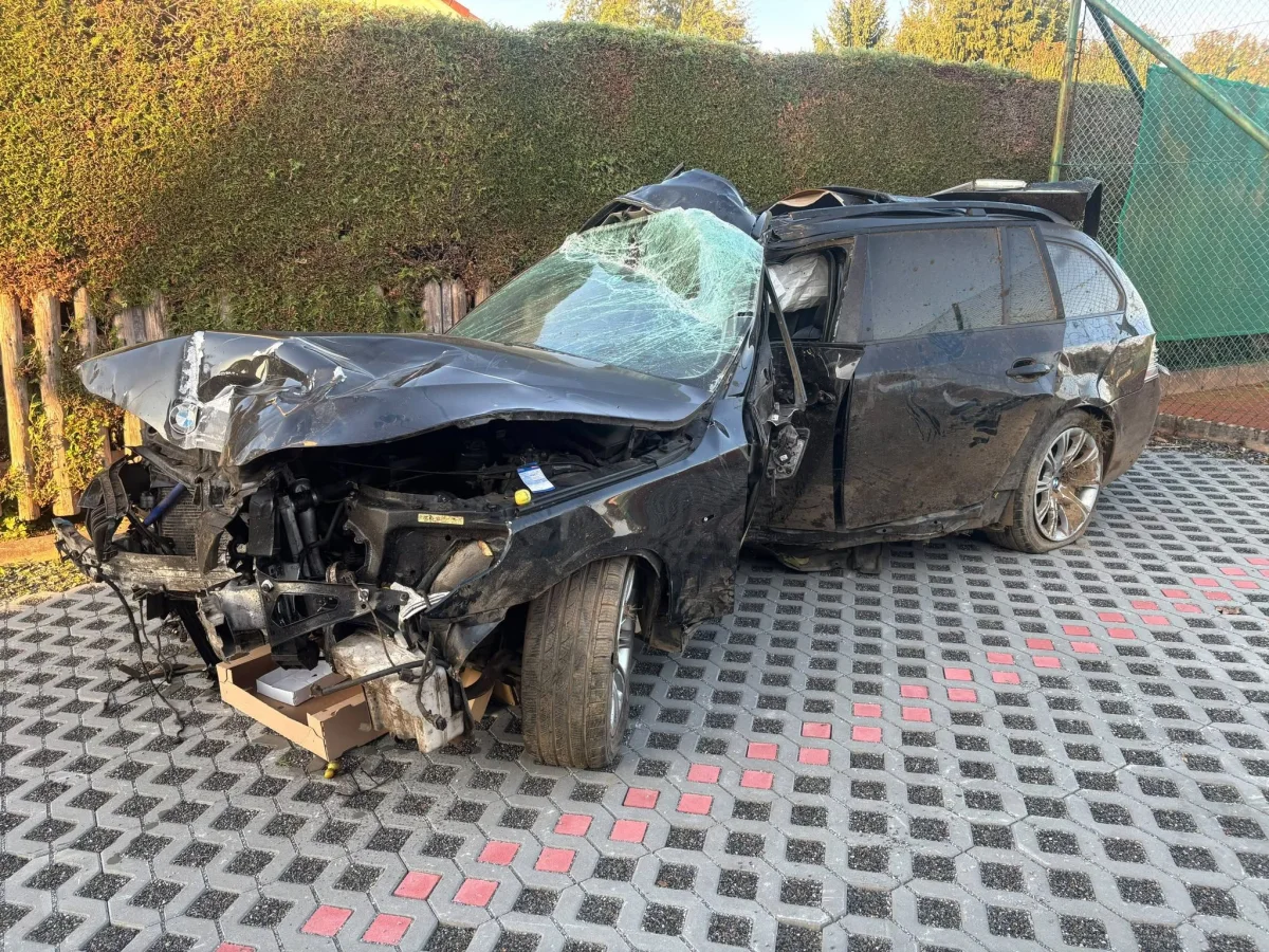Ein schwarzes Auto mit schwerer Front- und Dachschädigung steht auf gepflastertem Boden. Die Umgebung zeigt eine geschnittene Hecke.